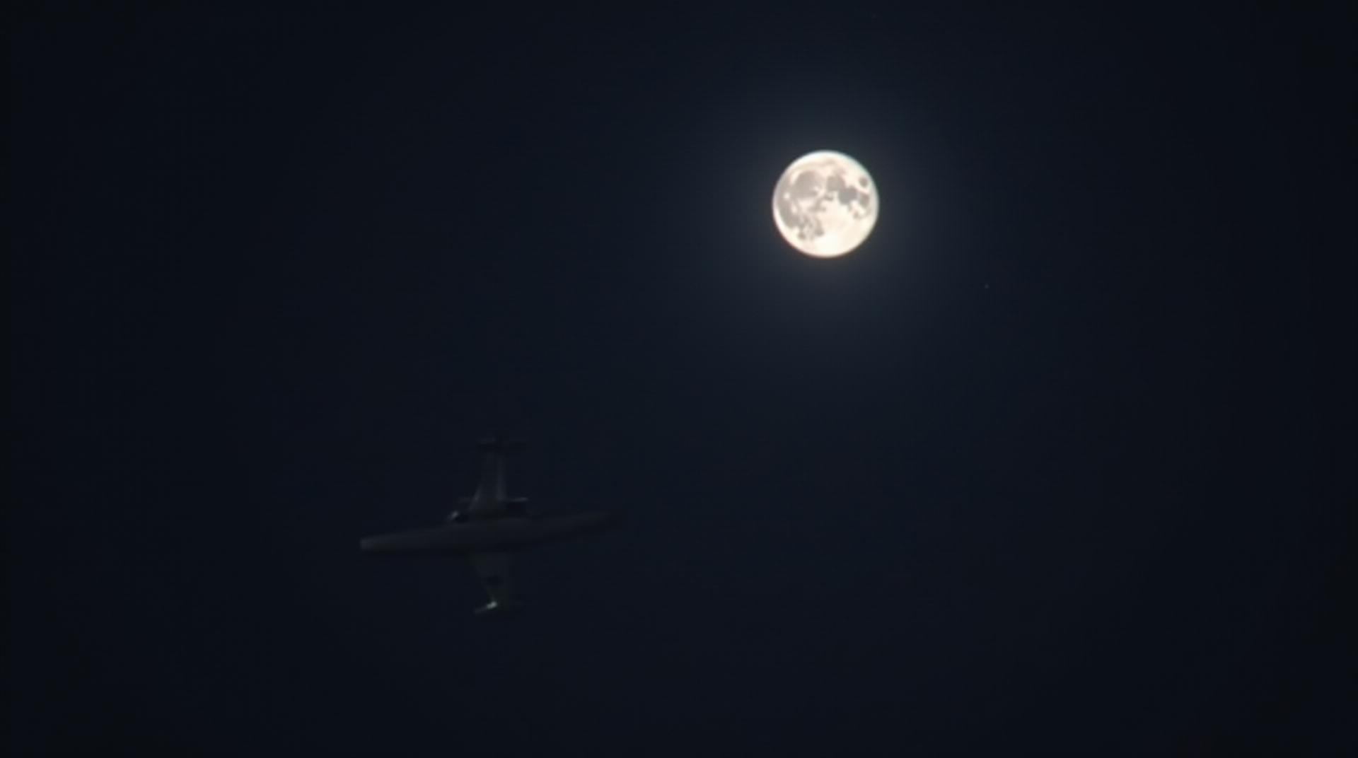 A plane is flying in the night sky, the moonlight is shining on the plane, and there is a bright moon in the distance